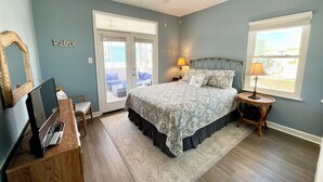 Una camera da letto con un letto, una scrivania con un portatile, una sedia, un ventilatore a soffitto e vista sulla spiaggia dalla finestra.