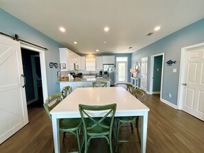 A spacious kitchen with white cabinets, a central island, and a dining area with a white table and green chairs. - Great views and next to boardwalk! (Gulf Shores)