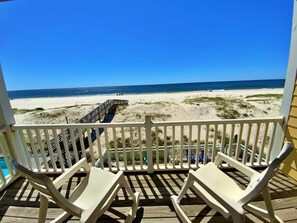 Une terrasse en bois avec des rampes blanches, deux chaises longues blanches et une vue sur la plage et l’océan.