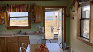 Uma cozinha com mesa de madeira, cadeiras e vista para a praia através de uma porta aberta.