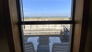 Vista de um quarto com uma grande janela para a praia. Há duas cadeiras de plástico brancas e uma mesa pequena na varanda.