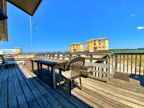 Una terraza de madera con una mesa y sillas, con vista a una playa y edificios al fondo.