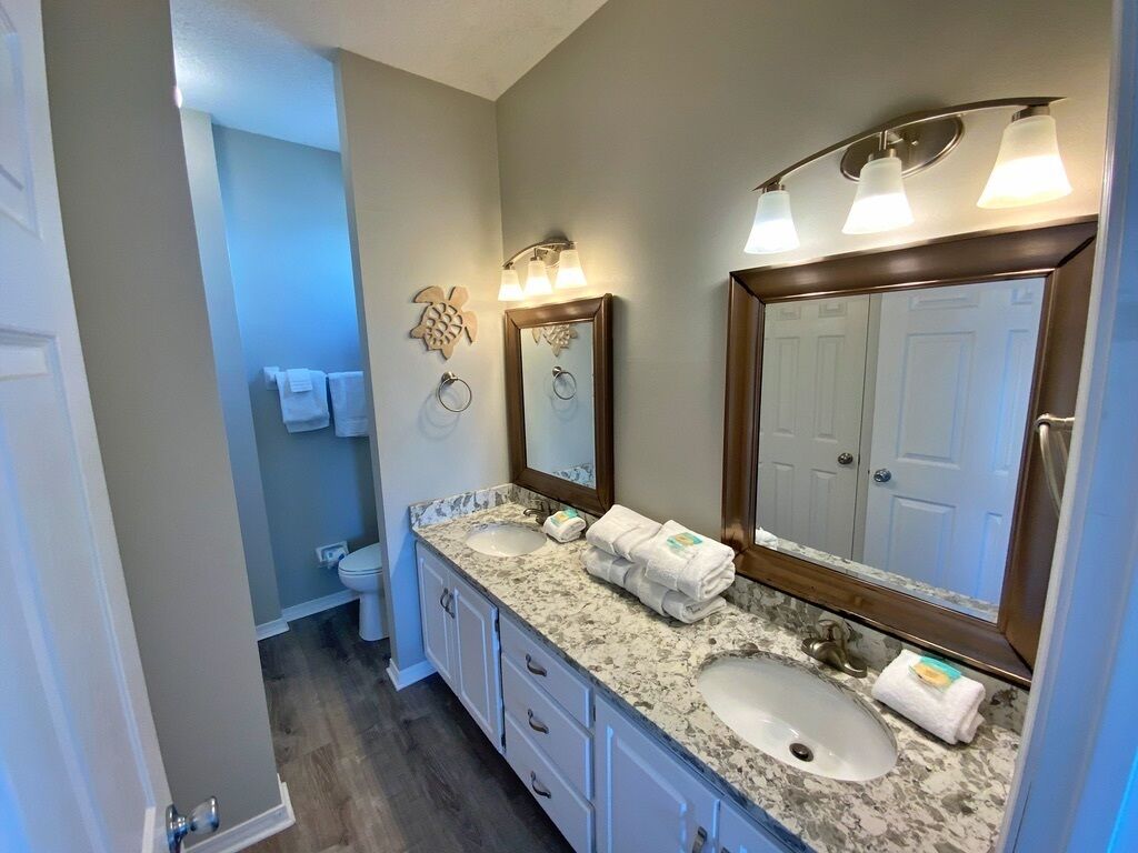Un baño con dos lavamanos, un espejo grande y apliques de pared. Se ve un inodoro al fondo.