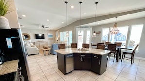 A modern kitchen with a central island, a dining area with wooden chairs, and a living room with a sofa and a TV. - Easy Beach Access & Pools!  Morgantown (Gulf Shores)