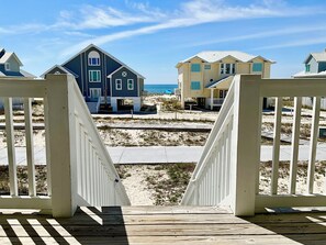 Eine Holzterrasse führt zu einem Anwesen am Strand mit zwei Häusern, einem blauen und einem gelben, und einem sandigen Strand im Hintergrund.