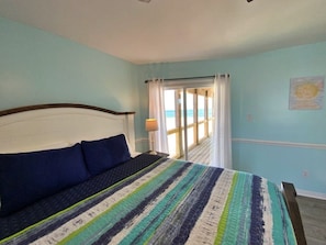 A bedroom with a bed, a view of the beach through a window, and a painting on the wall.