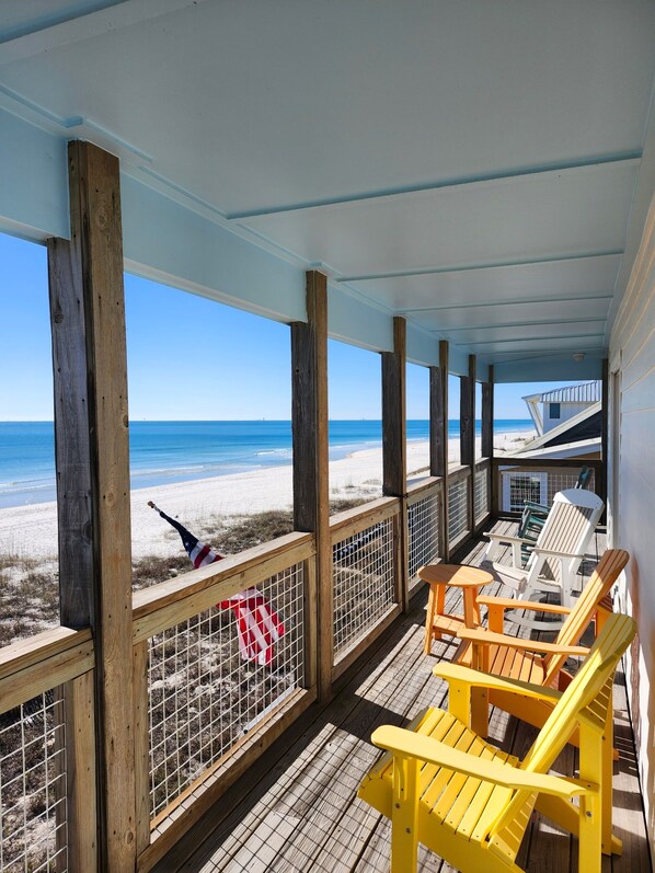 A covered wooden deck with a view of the beach, featuring wooden chairs and a table.