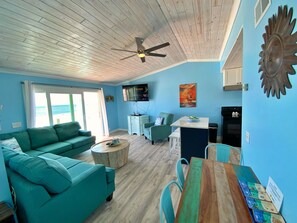 A living room with a teal sofa, a wooden coffee table, a dining table with chairs, a ceiling fan, a TV, and a painting on the wall. - Beachfront & Dog Friendly! (Gulf Shores)