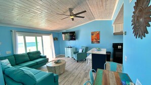 A living room with a teal sofa, a wooden coffee table, a dining table with chairs, a ceiling fan, a TV, and a painting on the wall. - Beachfront & Dog Friendly! (Gulf Shores)
