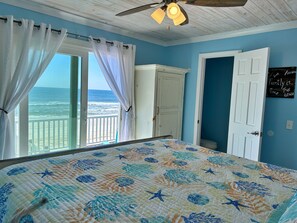 A bedroom with a view of the ocean, a bed with a floral quilt, a ceiling fan, and a door with a family-themed sign. - Beachfront & Dog Friendly! (Gulf Shores)