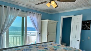 A bedroom with a view of the ocean, a bed with a floral quilt, a ceiling fan, and a door with a family-themed sign. - Beachfront & Dog Friendly! (Gulf Shores)