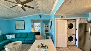 A living room with a teal sofa, a coffee table with a decorative bowl, a ceiling fan, and a kitchen area with a washing machine and refrigerator. - Beachfront in Fort Morgan - Dogs OK (Gulf Shores)