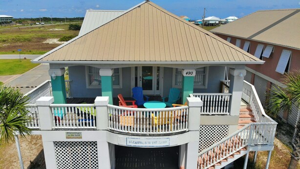 Une maison située en bord de mer, dotée d’un porche couvert, de meubles d’extérieur et d’une pancarte sur laquelle on peut lire : « Que le bonheur vous accompagne sur le sable ».