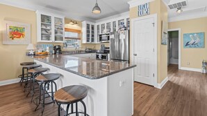 Una cocina con armarios blancos, encimera de granito y refrigerador de acero inoxidable. Hay cuatro taburetes en la isla, y un cuadro de un cangrejo en la pared.