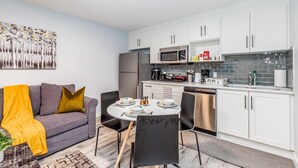 A modern kitchen with white cabinets, a dining table with chairs, a gray sofa with a yellow throw, and a painting on the wall. - Deluxe Suite w/Fast WiFi, Perfect for Long Stays (Airdrie)