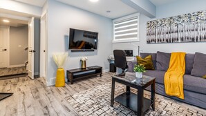 A modern living room with a flat-screen TV, a sofa, a coffee table, and a desk with a computer. - Deluxe Suite w/Fast WiFi, Perfect for Long Stays (Airdrie)