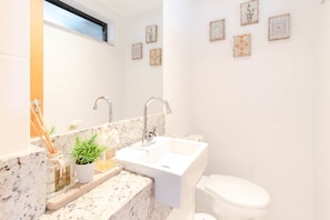 A bathroom with a white sink, two faucets, a mirror, and a toilet. There are decorative items on the sink counter, including a potted plant and a small bottle.