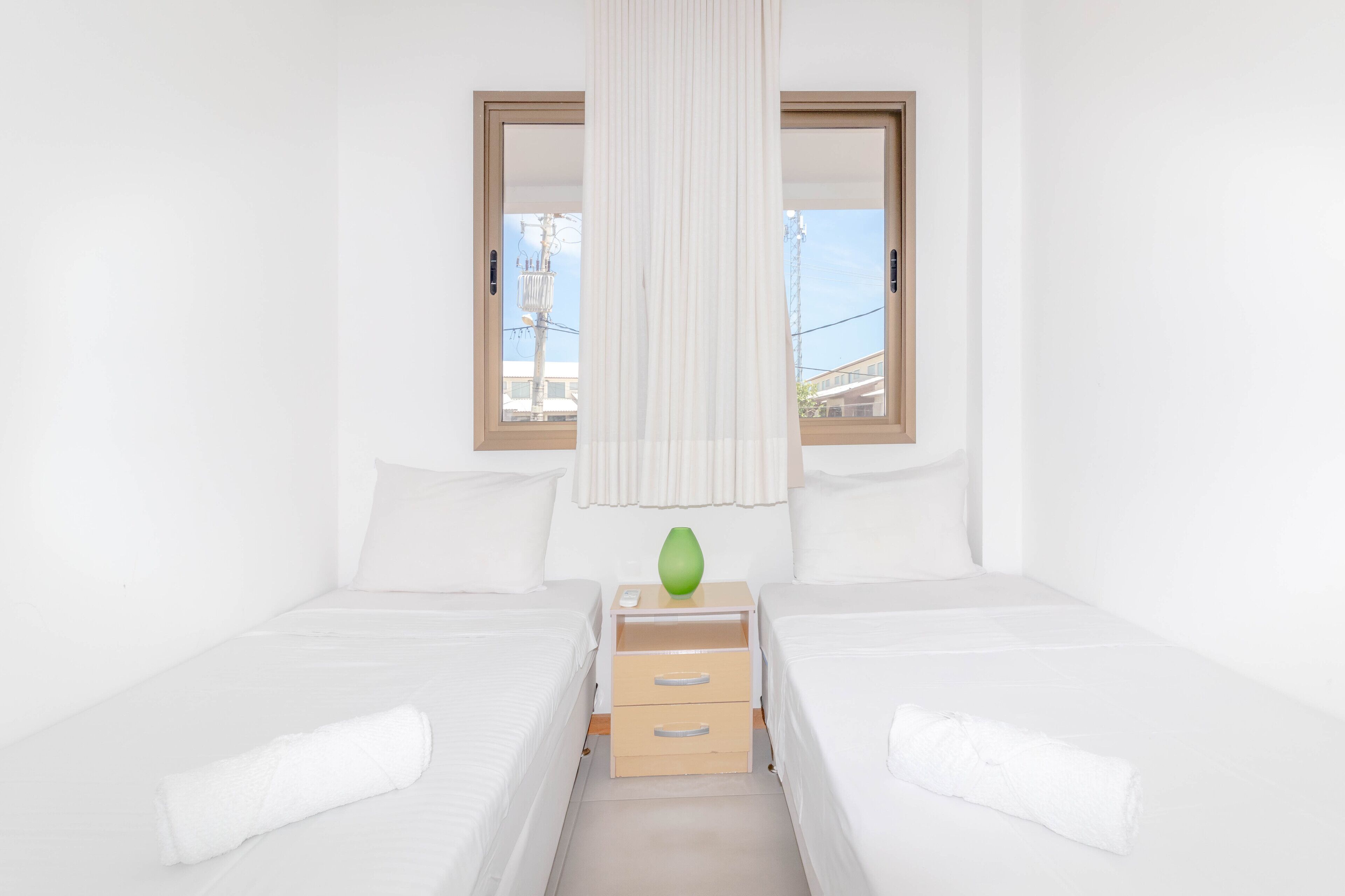Quarto de hotel com duas camas, uma mesa de cabeceira com gavetas, um vaso verde e uma janela com cortinas.