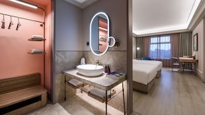 Une chambre d’hôtel moderne avec un grand miroir ovale, un lavabo rond, un banc en bois, un lit et un bureau avec une chaise.