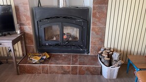 A wood-burning stove with a glass door, a television on a wooden stand, and a table with a chair.
