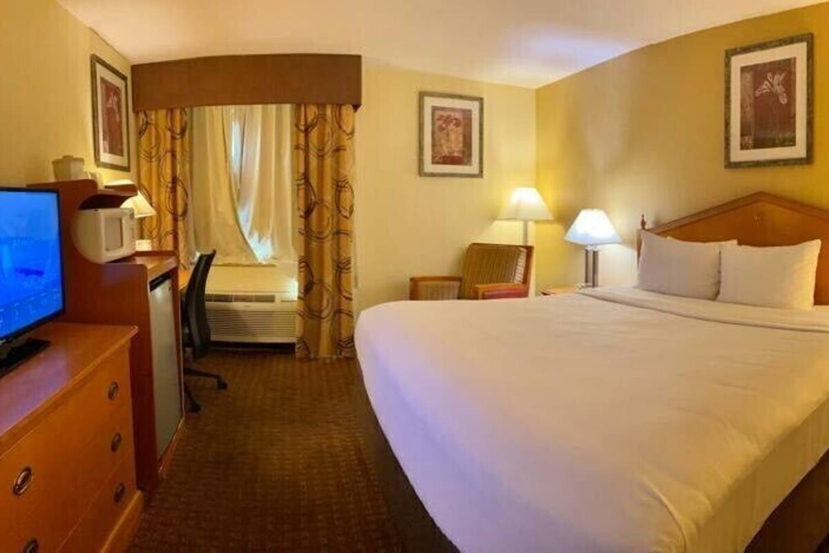 A hotel room with a bed, two bedside tables with lamps, a desk with a computer, a chair, and a television.