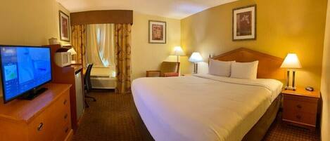 Una habitación de hotel con una cama grande, dos mesitas de noche con lámparas, un escritorio con ordenador, un televisor y una ventana con cortinas.