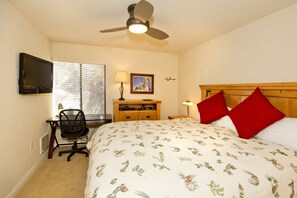 Une chambre à coucher comprenant un lit, un bureau, une chaise, une télévision, un ventilateur de plafond et une fenêtre avec des stores.