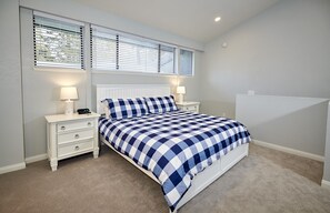 Une chambre à coucher comprenant un lit, deux tables de chevet, une commode, une fenêtre avec des stores et un luminaire au plafond.