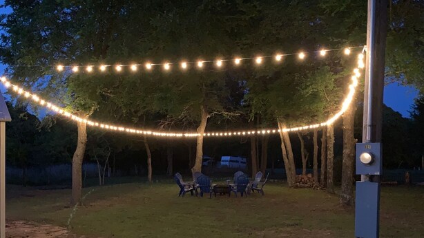 An outdoor seating area with string lights, surrounded by trees and a grassy lawn.