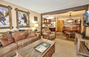A cozy living room with a sofa, armchairs, and a coffee table. There is a kitchen in the background with a dining area.