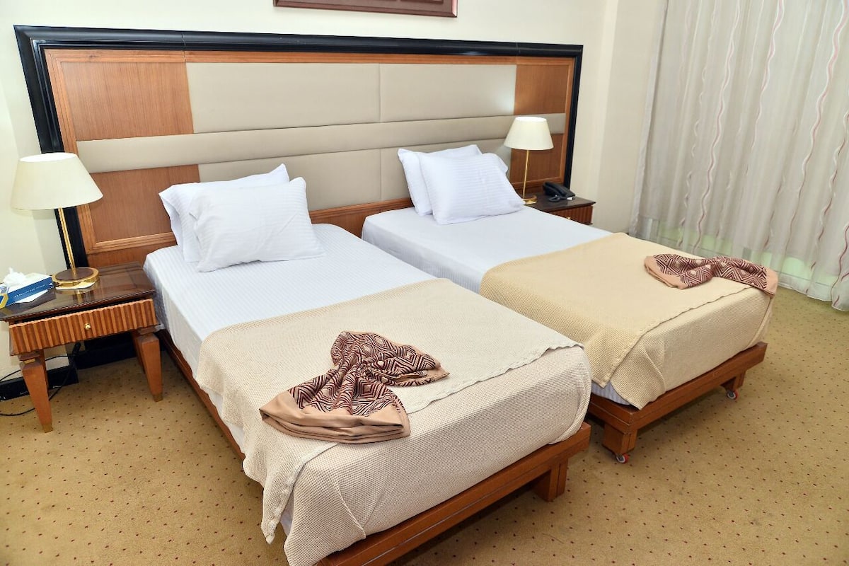 Quarto de hotel com duas camas, uma cabeceira de madeira, uma mesa de cabeceira com abajur e uma cortina.