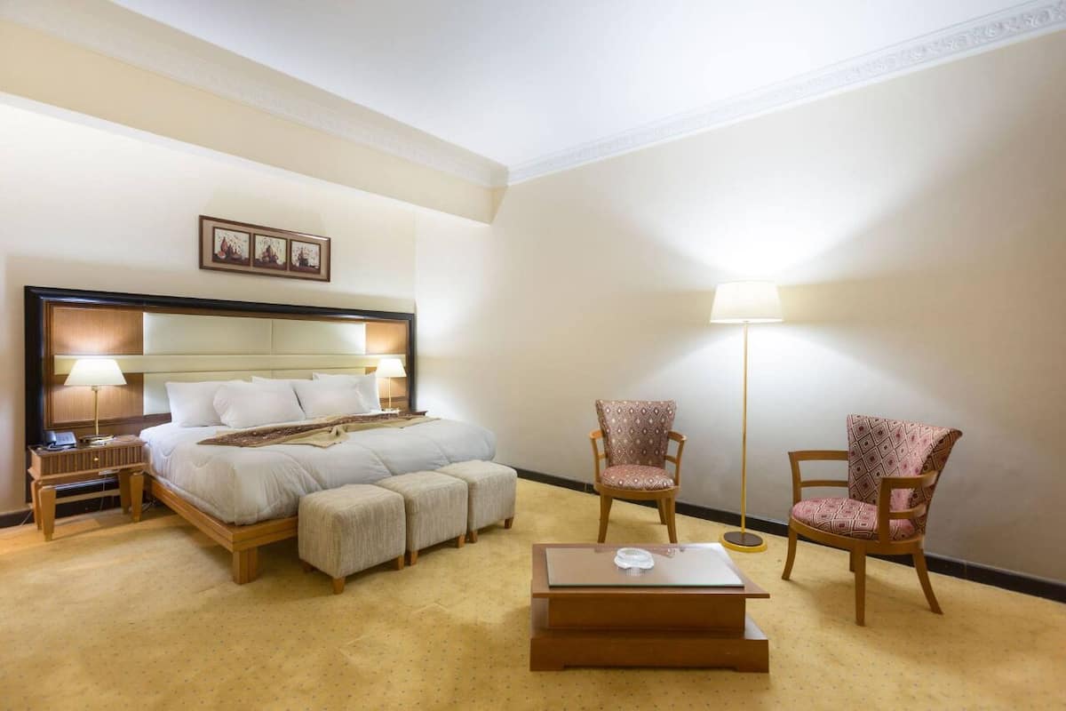 Quarto de hotel com uma cama, duas poltronas, uma mesa de cabeceira, um abajur e uma mesinha de centro.