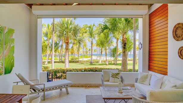 Una stanza con una grande finestra vista su un giardino tropicale e una spiaggia. Si vede un divano bianco, un tavolo di legno e una poltrona bianca.