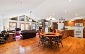 A spacious living room with a wooden floor, a dining area with a round table and chairs, and a kitchen with wooden cabinets and white appliances.