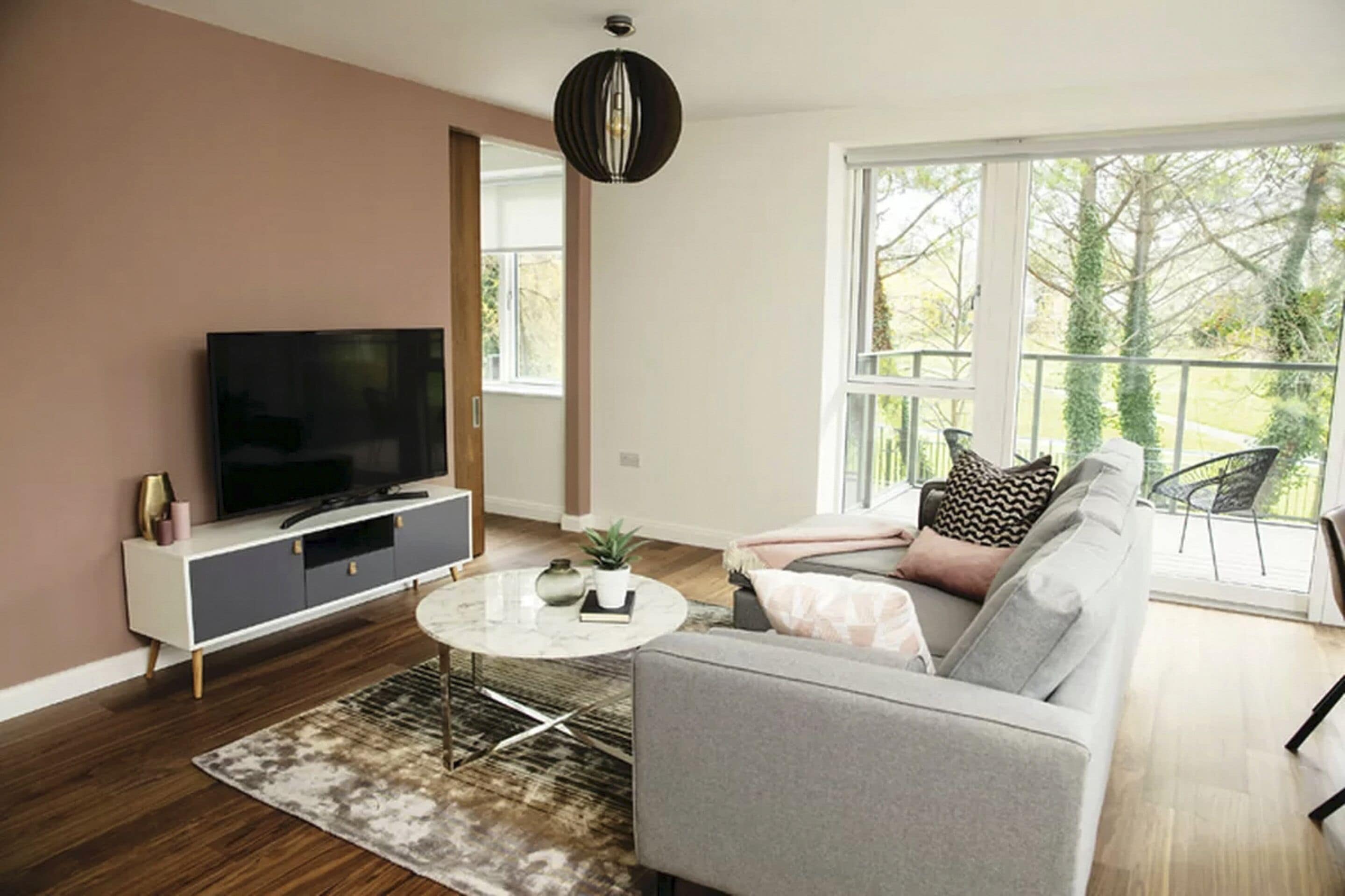 Ein modernes Wohnzimmer mit Fernseher, einer Couch, einem Couchtisch und einem großen Fenster mit Blick auf Bäume.