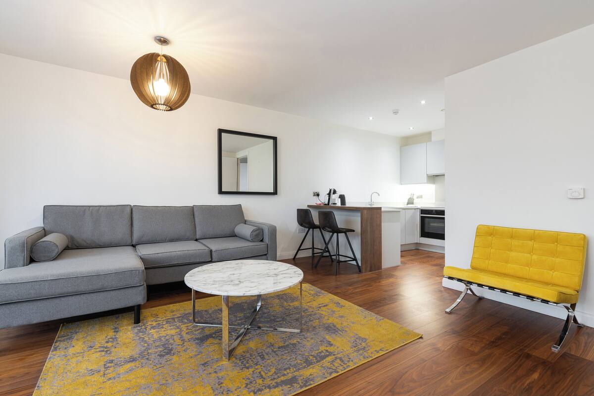 A modern living room with a gray sectional sofa, a small round table, and a yellow bench. A kitchen area is visible in the background with a bar counter and chairs.