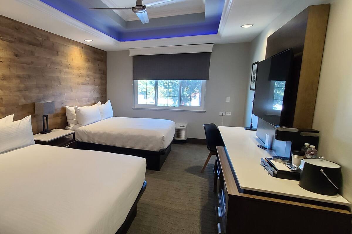 A hotel room with two beds, a desk with a flat-screen TV, a ceiling fan, and a window with a view of trees.