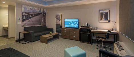 Habitación de hotel con sofá, una mesa de centro, televisión, escritorio y una silla, y un ventanal grande.