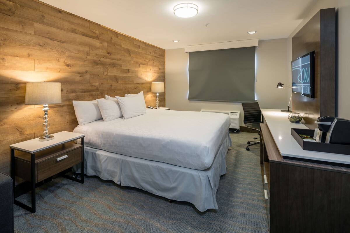 A hotel room with a large bed, wooden headboard, two bedside tables with lamps, a desk with a computer, and a TV on the wall.