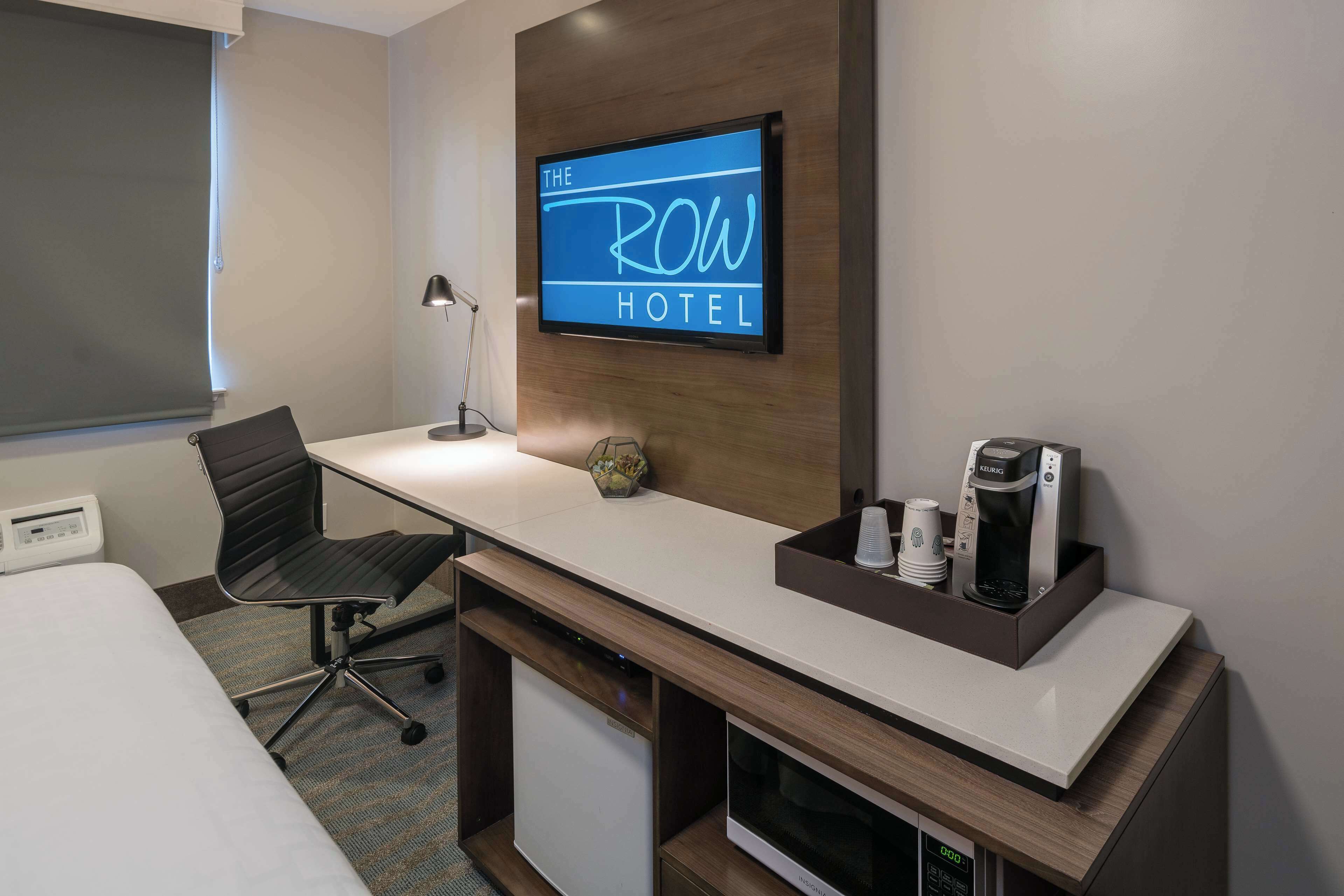 Une chambre d’hôtel équipée d’un lit, d’un bureau avec un ordinateur, d’une chaise, d’une machine à café et d’un téléviseur affichant le texte « ROW HOTEL ».