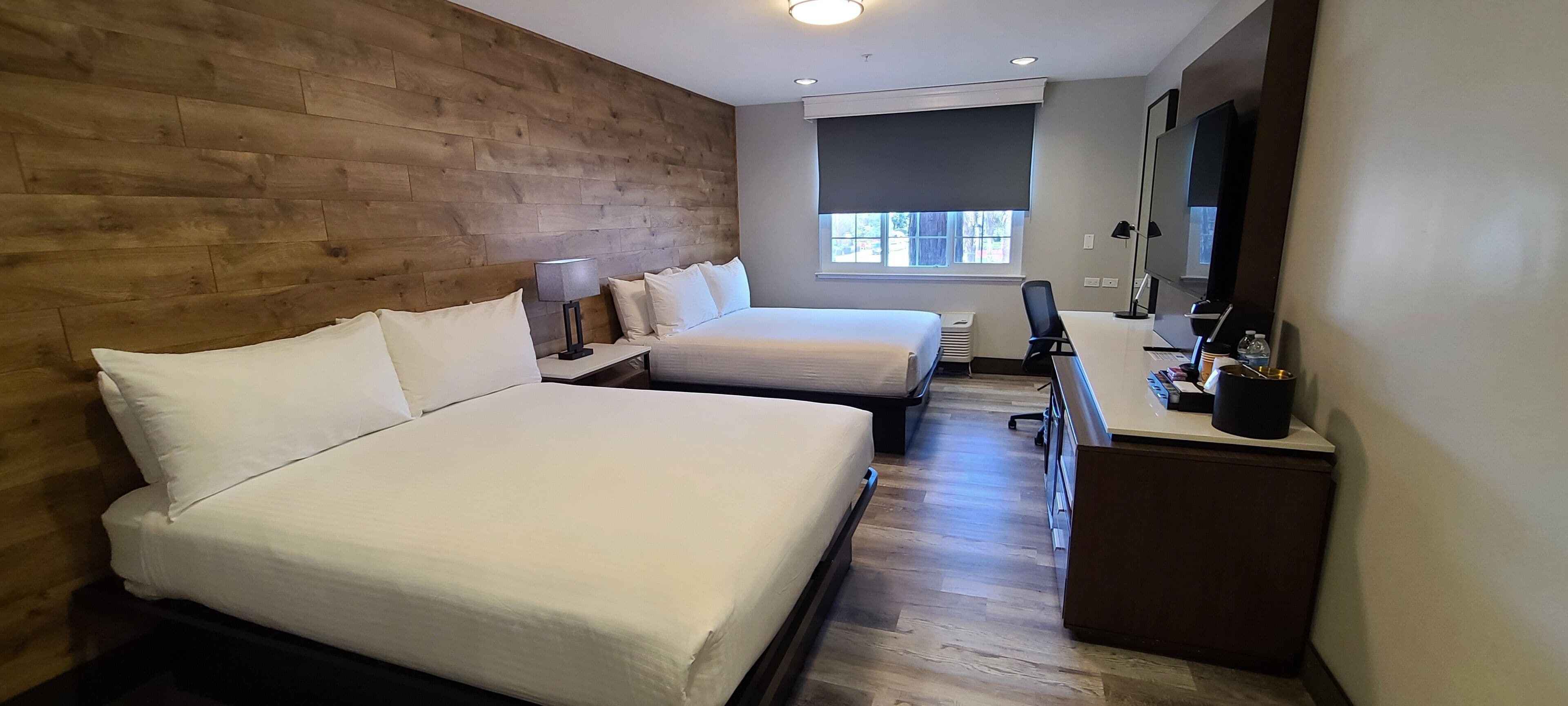 A hotel room with two beds, a desk with a computer, a TV, and a window with blinds.