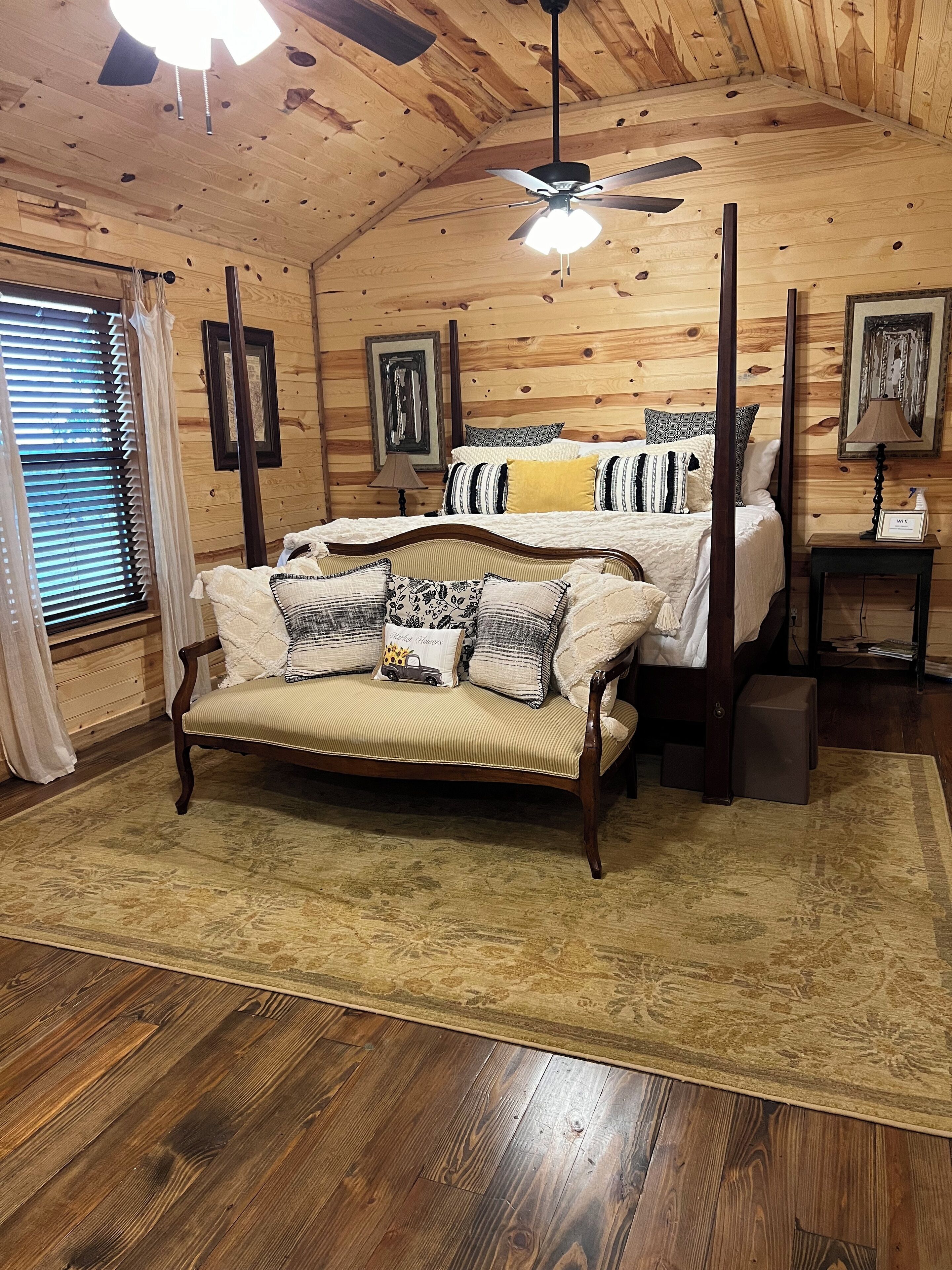 A cozy bedroom with a wooden ceiling, a bed with patterned bedding, a wooden chair, and a bedside table with a lamp.
