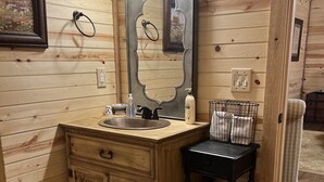 Un bagno in una cabina di legno con un mobile bagno in legno, un lavandino, uno specchio e un piccolo tavolo con articoli da toilette.