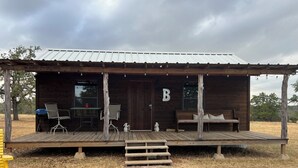 Una baita rustica con un portico, una panca di legno, un tavolo e la lettera 'B' sulla porta.