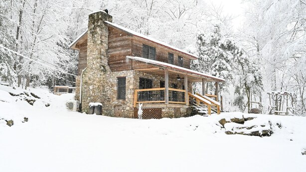 Una cabaña de troncos con chimenea de piedra, rodeada de árboles cubiertos de nieve.