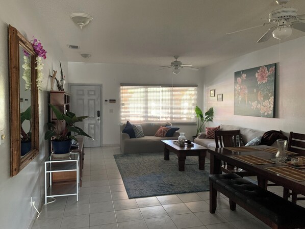 A spacious living room with a sofa, coffee table, and dining table. There is a large window with blinds, a ceiling fan, and a painting on the wall.