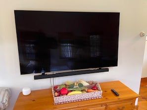 A flat-screen TV mounted on a wall above a wooden table with a fruit basket and a remote control.
