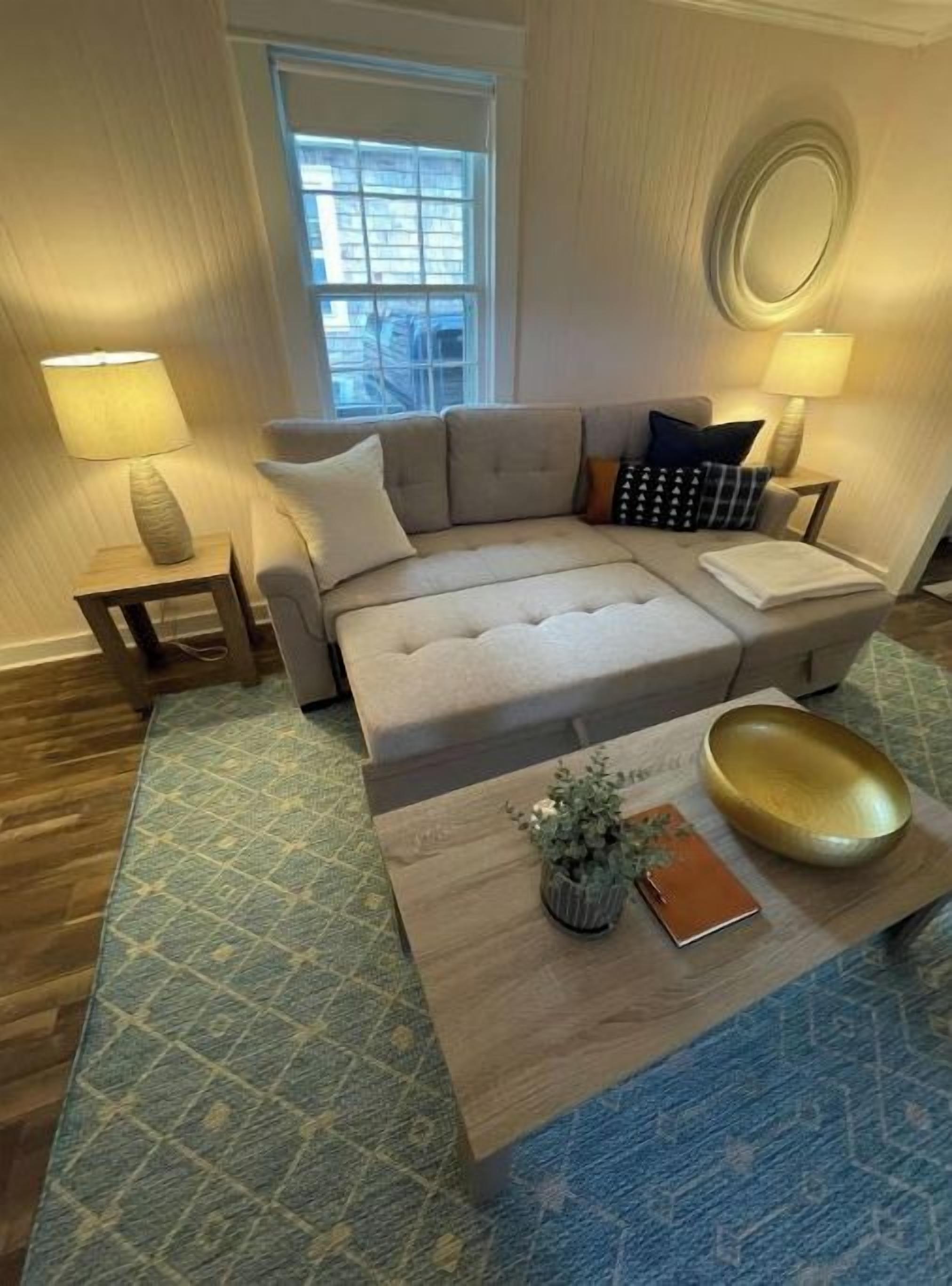 A living room with a beige sectional sofa, two side tables with lamps, a coffee table with a plant and a notebook, and a patterned rug.