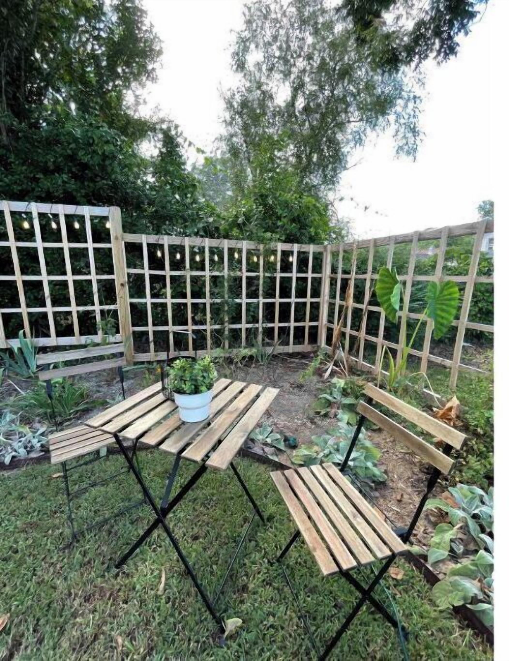 Een houten bank en tafel staan in een tuin met een houten hek en diverse planten.