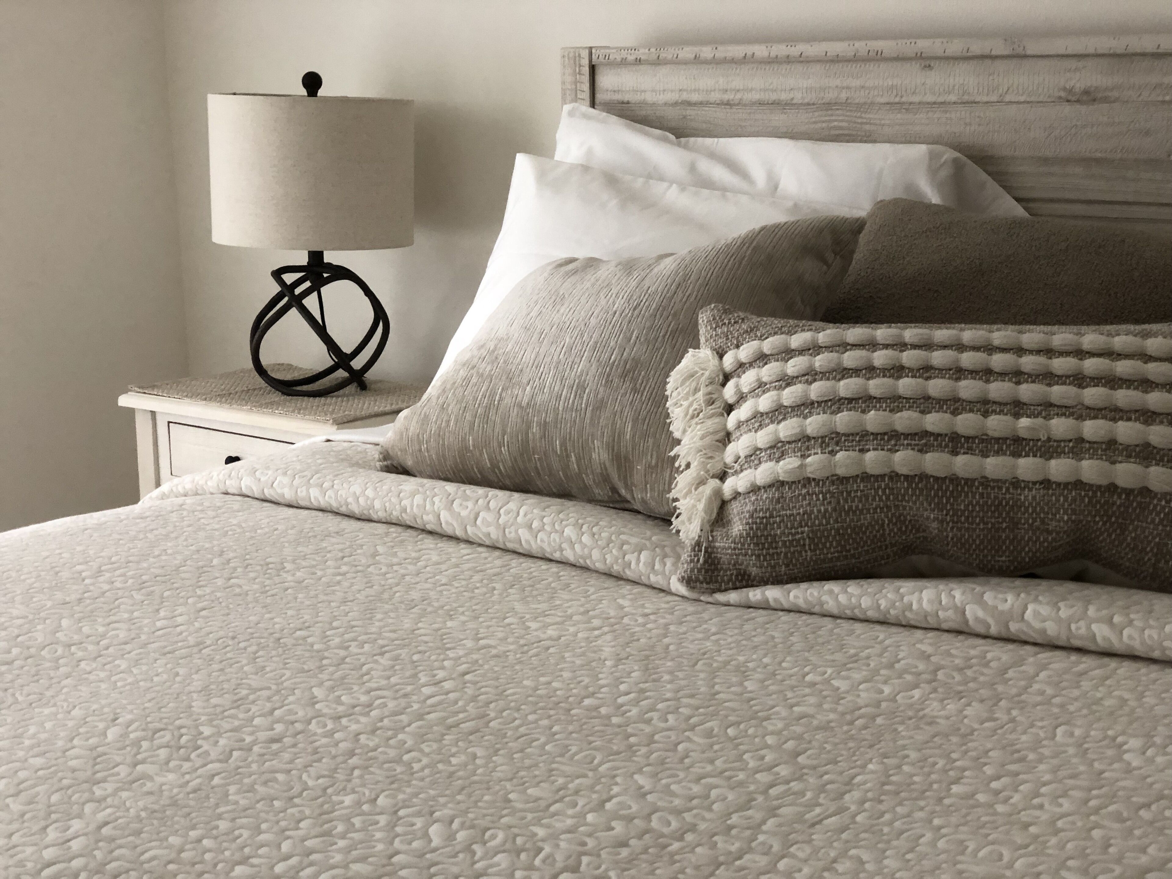 Cama bem arrumada com manta texturizada e várias almofadas, luminária de cabeceira e cabeceira de madeira.